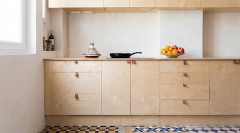 Cocina moderna con gabinetes de madera clara, encimera despejada, tajín, sartén y frutero con cítricos. Azulejos estampados en el suelo. Ventana a la izquierda.