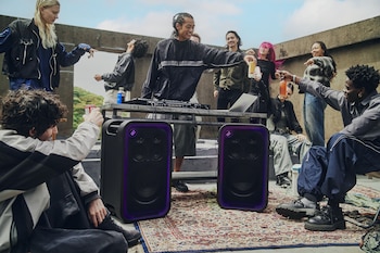 Jóvenes en una terraza junto a una mesa con equipo de música, una computadora y dos altavoces negros con bordes morados