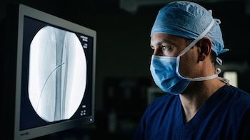 Un cirujano con mascarilla, gorro y traje azul contempla una radiografía de un hueso en una pantalla