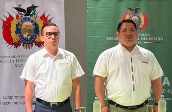 Dos hombres con camisas blancas con el logotipo de la Fiscalía posan frente a dos pancartas con el escudo de Bolivia
