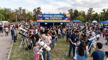 Un grupo de personas hace fila en un parque para mirar por diferentes telescopios junto a un cartel de la Semana Mundial del Espacio.