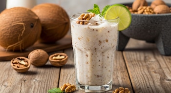 Un vaso de vidrio con agua fresca de coco y nuez, decorado con una rodaja de lima y menta, sobre una mesa de madera. Se ven cocos y nueces alrededor.