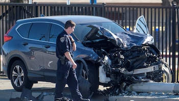 Oficial de policía examina un SUV compacto gris oscuro con la parte delantera severamente destrozada y escombros en el suelo. Una valla negra está detrás