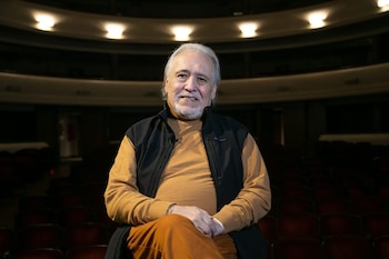 Pocho Sosa, un hombre de cabello y barba blancos, sonríe sentado en un teatro con filas de asientos rojos y luces en el techo, lleva cuello alto mostaza y chaleco negro