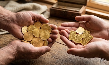 Primer plano de dos manos de un hombre; la izquierda sostiene una pila de monedas de oro y la derecha varios lingotes de oro sobre una mesa de madera