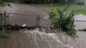 Vista de una inundación donde el agua turbia y con espuma fluye a través y sobre un cercado de malla metálica con un letrero de 'PRIVATE PROPERTY'. Se ven árboles y vegetación a los lados.