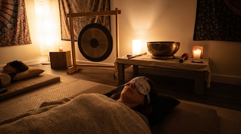 Una mujer con antifaz y cubierta con una manta, recostada sobre una colchoneta. Al fondo, un gong, un cuenco tibetano y luz tenue de velas.