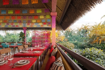 Mesas con manteles rojos, vajilla y copas bajo un techo de paja con papel picado, junto a una barandilla frente a un río rodeado de árboles