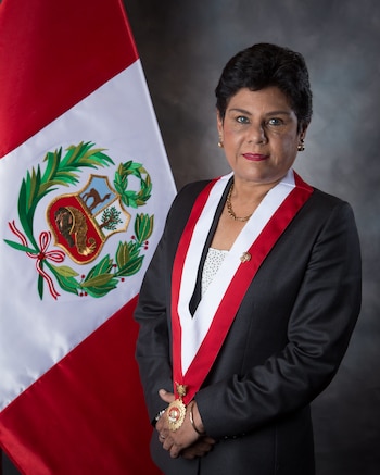 Retrato de mujer de cabello oscuro, traje negro, camisa de puntos, collar y aros dorados. Lleva una banda presidencial con medalla. Detrás, la bandera de Perú