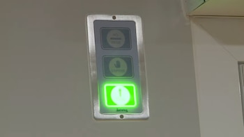 Panel de control plateado montado en pared blanca, con tres indicadores. Los superiores dicen "NO PASE" y "ESPERE", y el inferior es verde brillante con un número 1