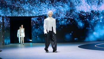 Robot humanoide con cabello blanco, camisa blanca, pantalón oscuro y guantes negros desfila en pasarela. Dos modelos humanas lo siguen. Fondo de pantalla digital azul con destellos
