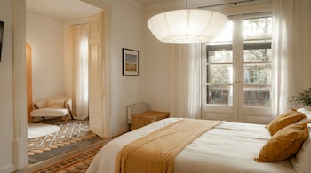 Un dormitorio luminoso con una cama blanca y una manta amarilla, una lámpara de papel redonda, una ventana grande con cortinas claras y un pasillo con un sillón.