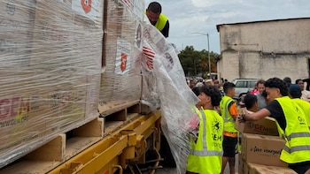 Palés de cajas de ayuda humanitaria envueltas en plástico sobre un camión, varias personas descargando y distribuyendo las cajas, y una multitud al fondo