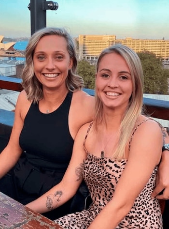 Dos mujeres sonriendo posan sentadas en una mesa al aire libre. La mujer de la izquierda lleva un top negro y la de la derecha un vestido estampado. Edificios de fondo