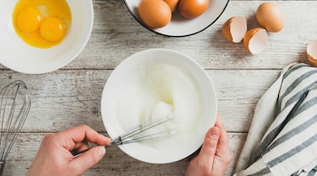Manos batiendo claras de huevo en un bol con un batidor de varillas. Yemas separadas, huevos enteros y cáscaras sobre una mesa de madera rústica.