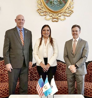 Dos hombres en traje y una mujer con blazer blanco posan de pie. Detrás, un sofá y un cuadro dorado. Sobre una mesa, banderas de EEUU, Guatemala y otra insignia