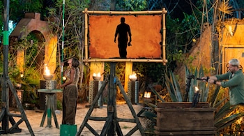 Dos personas en un escenario de juego nocturno con plantas, antorchas y estructuras de madera. Un cartel de bambú muestra una silueta oscura.