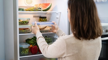 Persona de espaldas guarda un recipiente rectangular con pechugas de pollo cocidas y tapa azul en la heladera, que contiene sandía, espárragos y otros vegetales.