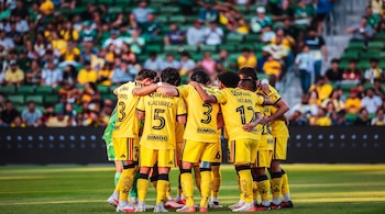Nueve jugadores de fútbol con uniformes amarillos, números y patrocinadores visibles, forman un círculo en el césped. Fondo de gradas con público.