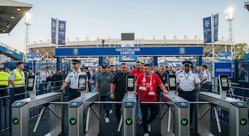 Entrada de un estadio de fútbol con tres torniquetes de reconocimiento facial activos, personal de seguridad y aficionados ingresando.
