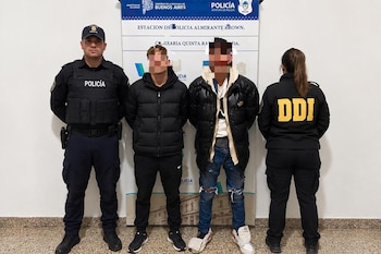 Dos hombres con los rostros cubiertos permanecen de pie entre dos efectivos policiales frente a una pancarta de la policía de Buenos Aires