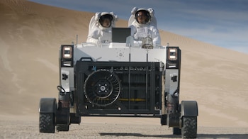 Vista frontal de dos astronautas con trajes espaciales blancos dentro de un vehículo explorador blanco y negro, conduciendo sobre arena con dunas al fondo bajo un cielo azul