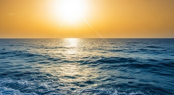 Vista panorámica del océano azul con olas. El sol brilla intensamente en el cielo naranja-amarillo, reflejándose en el agua y creando destellos luminosos.