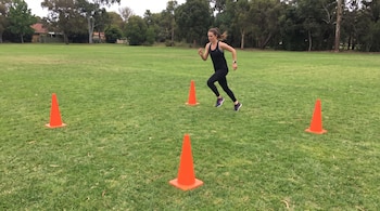 Mujer corriendo en un campo de hierba con cuatro conos naranjas. Viste ropa deportiva negra y reloj en la muñeca. Árboles en el fondo.