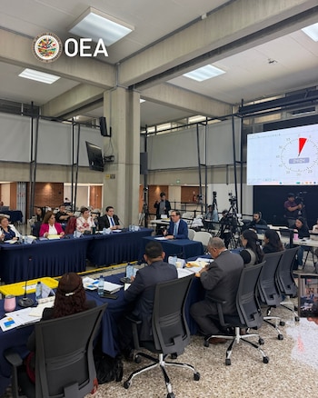 Varias personas sentadas a mesas con manteles azules participan en una reunión frente a una pantalla digital y cámaras sobre trípodes