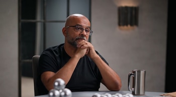 Mo Gawdat, hombre calvo con barba y gafas, camiseta negra, sentado en una mesa con manos cruzadas, mirando pensativo. En la mesa hay esferas metálicas y una taza