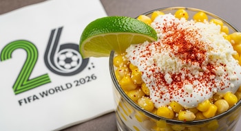 Vaso de esquites con maíz, crema, queso, chile en polvo y rodaja de limón. Al lado, una servilleta blanca con el logo estilizado FIFA World 2026.