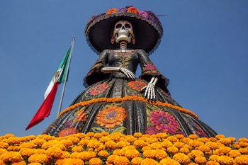 Figura de una catrina con sombrero y vestido oscuro rodeada de flores de cempasúchil, con la bandera de México y el cielo de fondo.