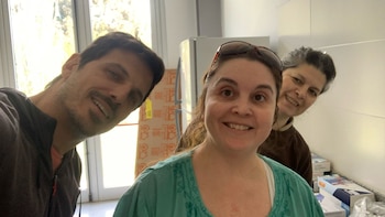 Tres personas sonríen a cámara en un interior. Un hombre a la izquierda y dos mujeres, una en el centro y otra a la derecha, aparecen en la imagen. Se ve una nevera y cajas en el fondo