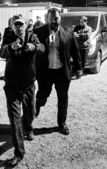 Fotografía en blanco y negro. Dos hombres caminan. Uno lleva gorra y suéter. El otro usa traje oscuro. Se ven un vehículo, estructuras y personas al fondo