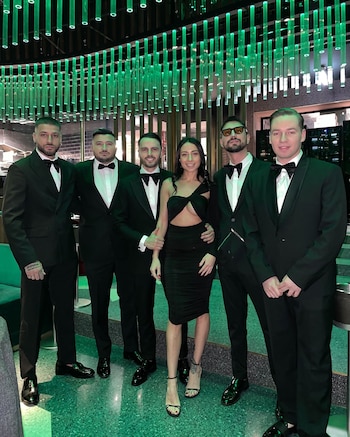 Cinco hombres con esmoquin y una mujer en vestido negro posan sonriendo en un salón con decoración de tubos luminosos verdes y piso de terrazo