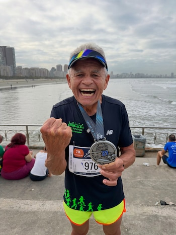 Un hombre mayor con visera y dorsal deportivo muestra una medalla de la Media Maratón de Santos frente al mar y la playa