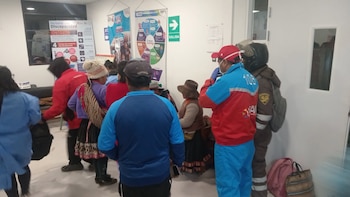 Grupo de personas, incluyendo mujeres con vestimenta tradicional y hombres con uniformes, en una sala con paredes blancas, carteles y un letrero de salida