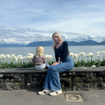 Una mujer y una niña pequeña están sentadas en un muro de piedra junto a flores blancas, con un gran lago y montañas de fondo