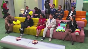 Once personas están en una sala: una mujer de cabello rojo de pie con micrófono, diez personas sentadas en sofás amarillo, rosado y naranja. Alfombra verde y mesa plateada.