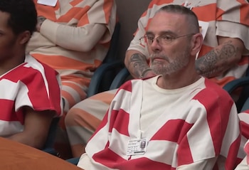 Hombre con barba, gafas y corte de pelo rapado, vestido con camisa a rayas rojas y blancas, y una placa de identificación, sentado junto a otros hombres con uniformes a rayas naranjas