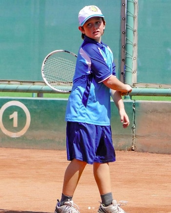 Un niño con gorra blanca y ropa deportiva azul en una cancha de tenis de arcilla, sosteniendo una raqueta y mirando hacia la derecha