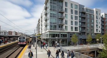 Vista exterior de un complejo de apartamentos moderno de varios pisos junto a una estación de tren con un tren en el andén y personas caminando. Cielo azul con nubes.
