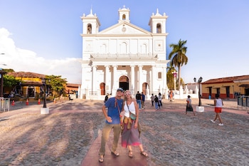 Un hombre y una mujer sonrientes posan frente a una gran iglesia colonial blanca con dos campanarios, en una plaza empedrada bajo un cielo azul claro