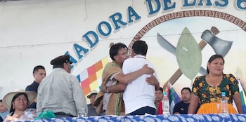 Dos hombres se abrazan en un estrado frente a un mural con un emblema y la bandera wiphala, acompañados por otras personas sentadas