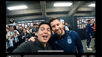 Selfie de dos hombres sonriendo a la cámara, uno es Lionel Messi y el otro un fan. De fondo se ven espectadores desenfocados y luces brillantes del estadio.