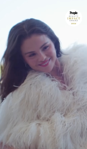 Selena Gomez apoya la mano derecha en la sien mientras viste una prenda cubierta de plumas blancas