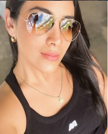 Primer plano de Lilia Moretti, mujer con cabello oscuro, gafas de sol aviador con reflejos de cielo, arete de aro dorado y collar fino