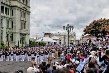 Una banda militar vestida de uniforme blanco y azul marcha en la calle ante una multitud en gradas que sostiene banderas