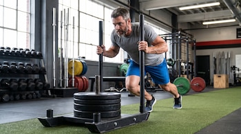 Um homem de 40 anos, de camiseta cinza e short azul, empurra um carrinho de musculação sobre um tapete verde na academia.