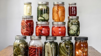 Un arreglo piramidal de doce tarros de vidrio llenos de alimentos encurtidos y conservas, incluyendo pepinos, zanahorias, pimientos y kimchi, sobre una superficie de madera clara.